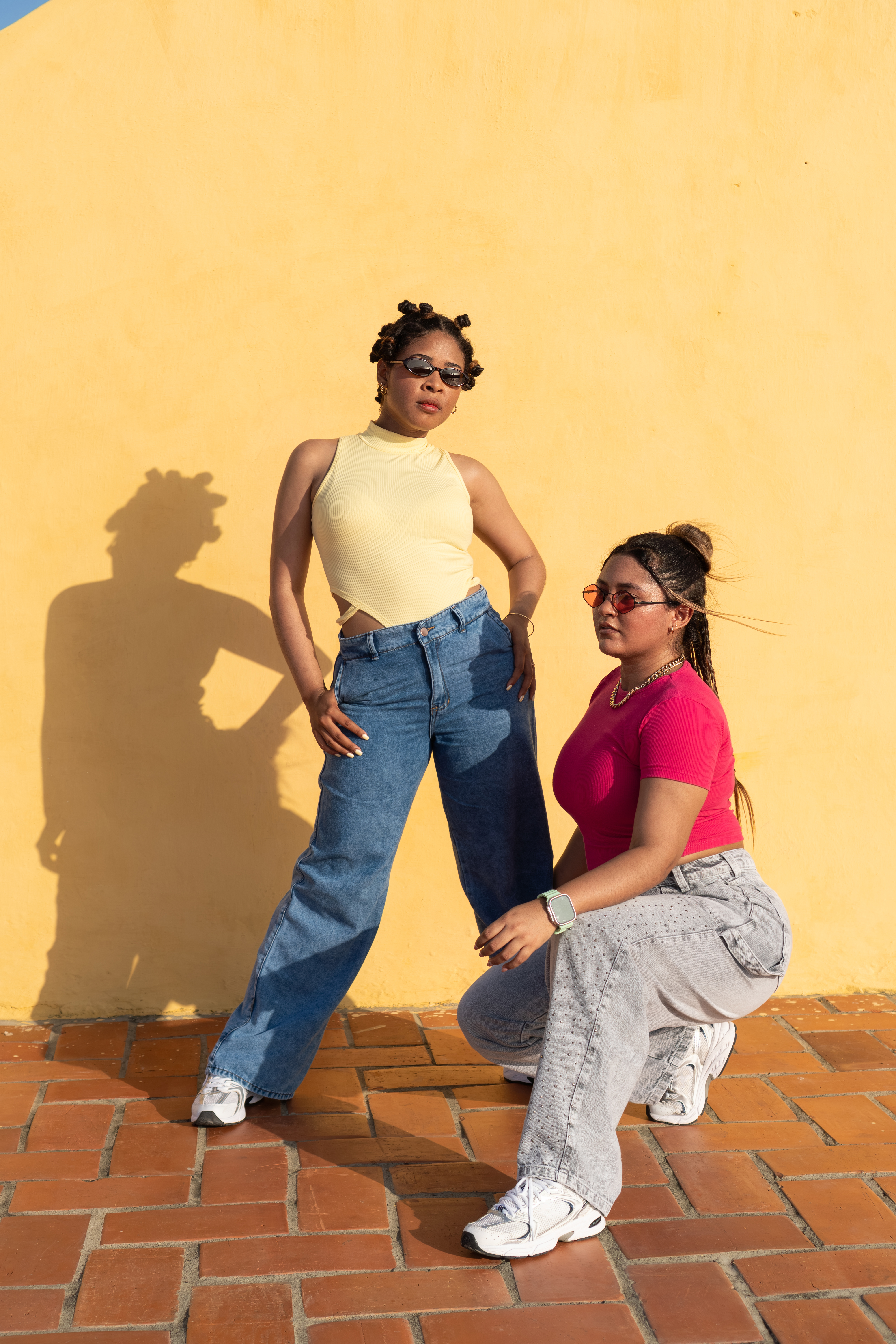 Two young adult women striking poses against a vibrant yellow wall in urban street fashion